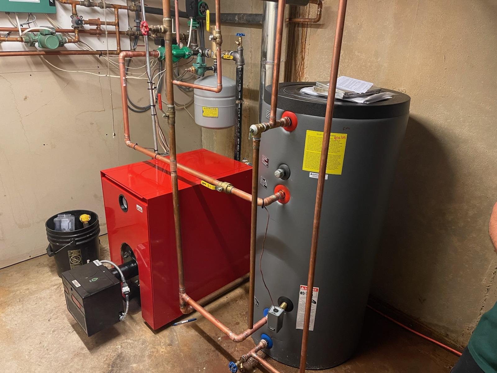 Side view of basement heating system featuring a bright red oil-fired boiler with a front-mounted burner, connected to an indirect white water heater tank and copper piping. The setup includes an expansion tank mounted above the unit and is installed against an unfinished wall with signs of past heat exposure.