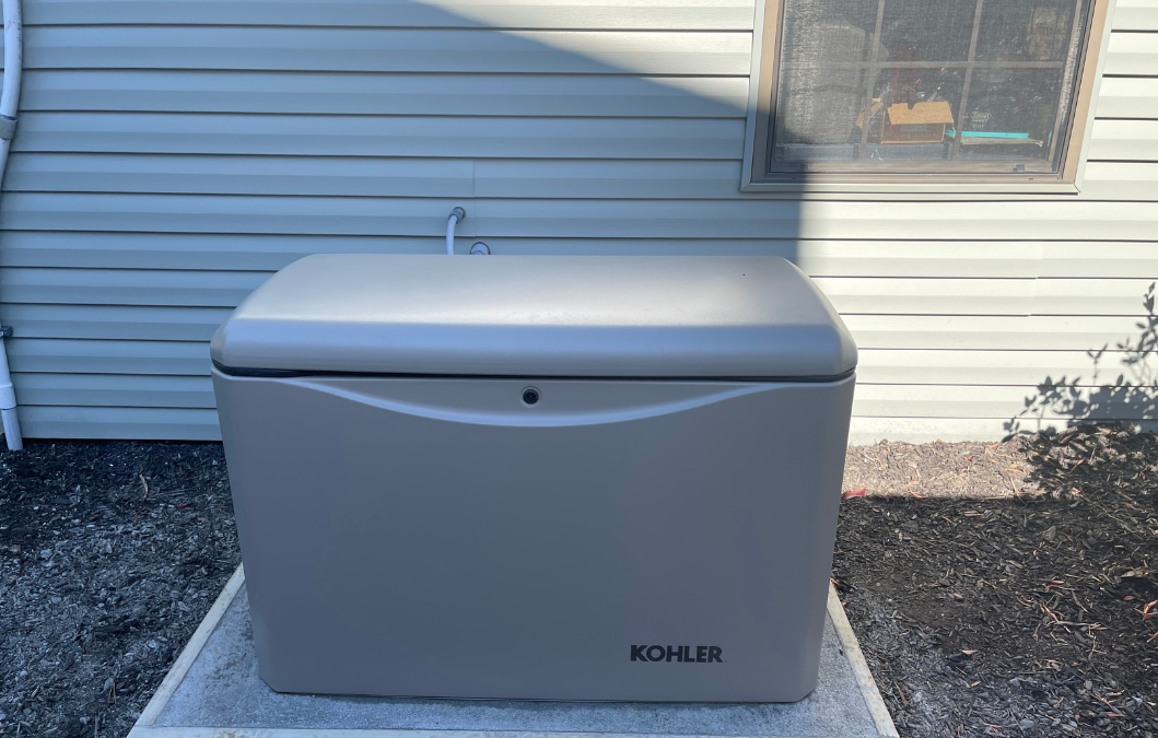 Outdoor standby generator by Kohler installed on a concrete pad next to a house with vinyl siding. The generator is positioned beneath a window and connected to utility lines, ready to provide backup power during outages.