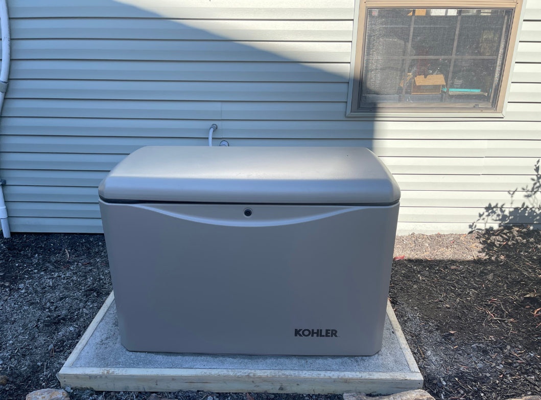 Outdoor standby generator by Kohler installed on a concrete pad next to a house with vinyl siding. The generator is positioned beneath a window and connected to utility lines, ready to provide backup power during outages.